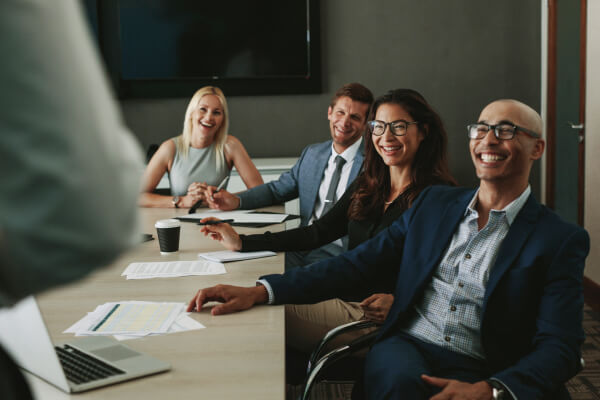 Four people in business attire sit in a conference room, smiling and laughing during a meeting. Papers and laptops are on the table in front of them, suggesting a professional setting. They appear engaged with discussions about HCM solutions, paying close attention to someone standing out of the frame.